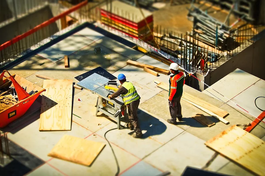 The photo shows two construction workers operating a table saw on a concrete foundation