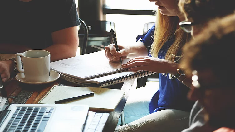 The photo shows a group of people working together in a workshop or meeting and taking notes.